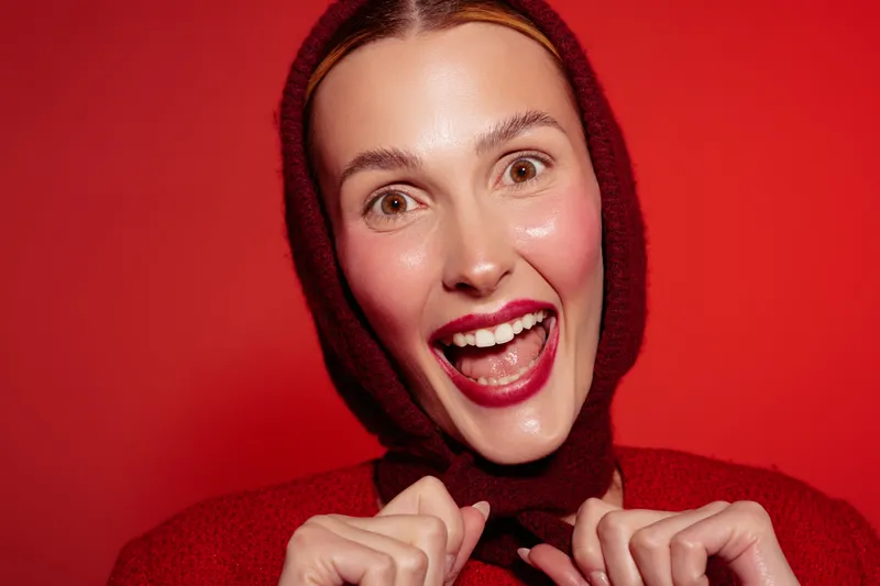 Happy woman in red sweater and hood smiles widely with bright lipstick on red background. Close-up portrait with joyful expression and glowing skin. Concept of happiness and fashion.