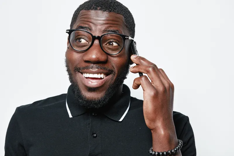Black man with dark skin and glasses smiling while talking on smartphone in casual black polo shirt against white background. Positive expression and communication concept.