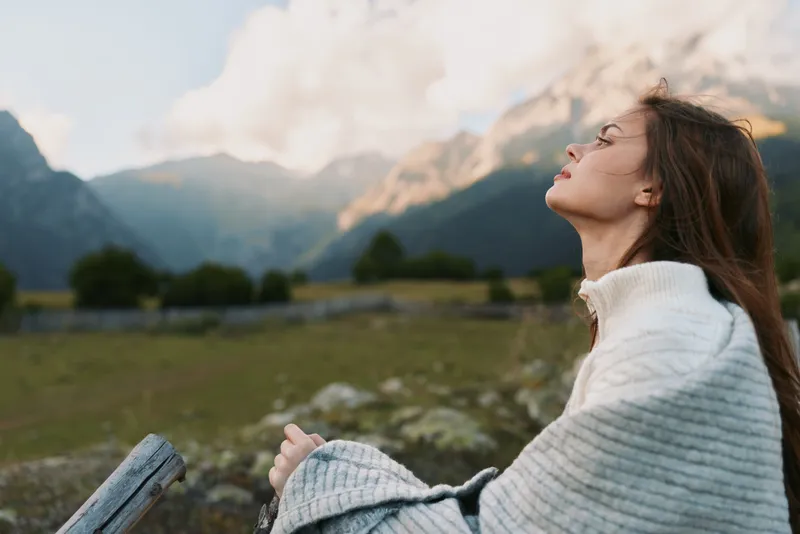 Young woman with long hair wearing a white sweater enjoys fresh mountain air with closed eyes in a peaceful outdoor setting during daytime.