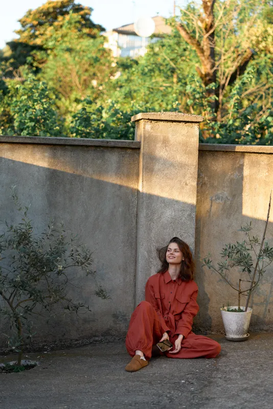 Young woman in a rust jumpsuit sits relaxed on the ground beside a sunlit courtyard wall, enjoying a quiet moment with a gentle smile, framed by warm natural light