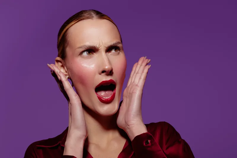 Surprised woman with red lipstick and slicked hair holds hands near face, looking shocked on purple background. Concept for emotion, reaction, and expression in portrait photography.