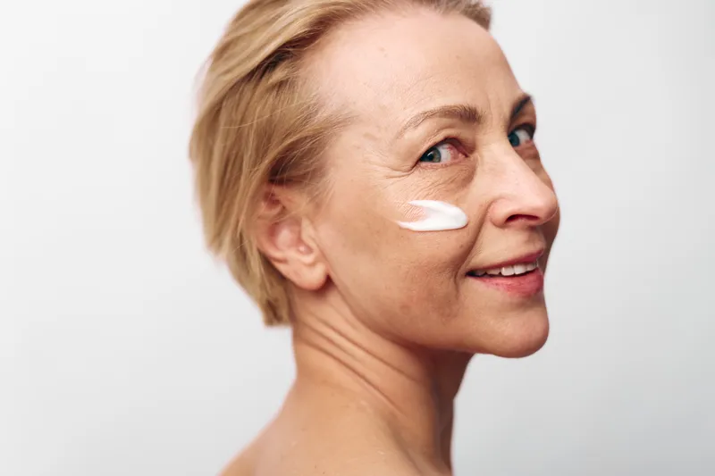 Mature woman with short blonde hair applies white face cream on cheek showing natural skin and smile against white background. Concept for skincare, aging beauty, self-care, and wellness.