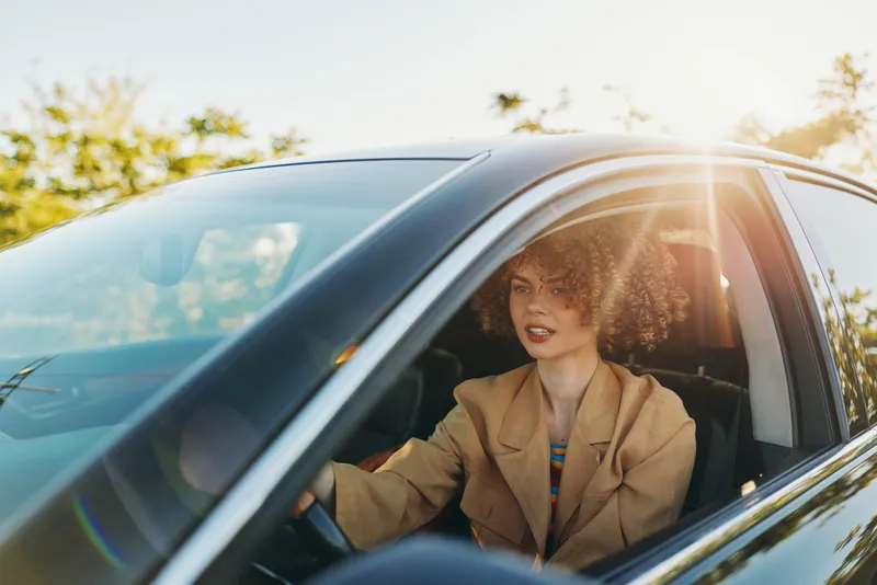 Young woman drives a car at golden hour, smiling softly as warm sunlight streams through the window, creating a relaxed nostalgic mood with a hint of retro film vibe