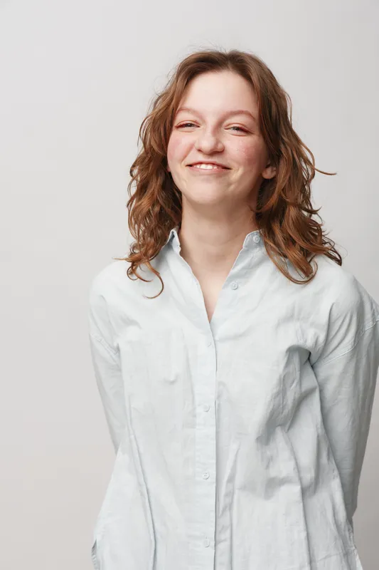 Smiling young woman with curly hair wearing a white shirt stands against a light background. She looks confident and happy, expressing natural beauty and casual style.
