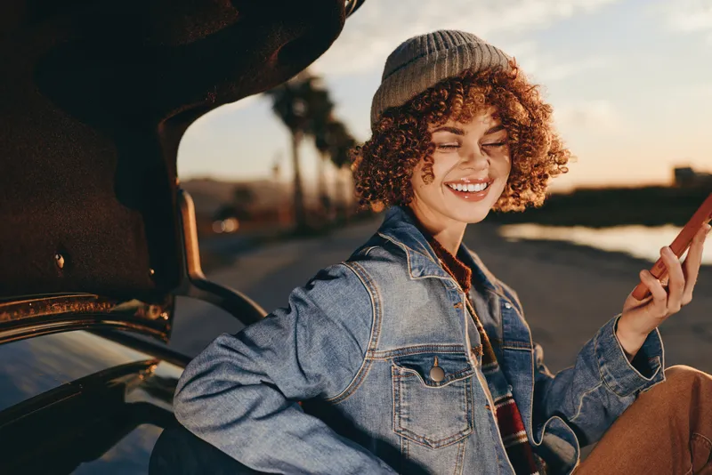 Woman lifestyle, smiling in a car at sunset, curly hair, denim jacket, peace sign gesture, warm road background, casual travel moment