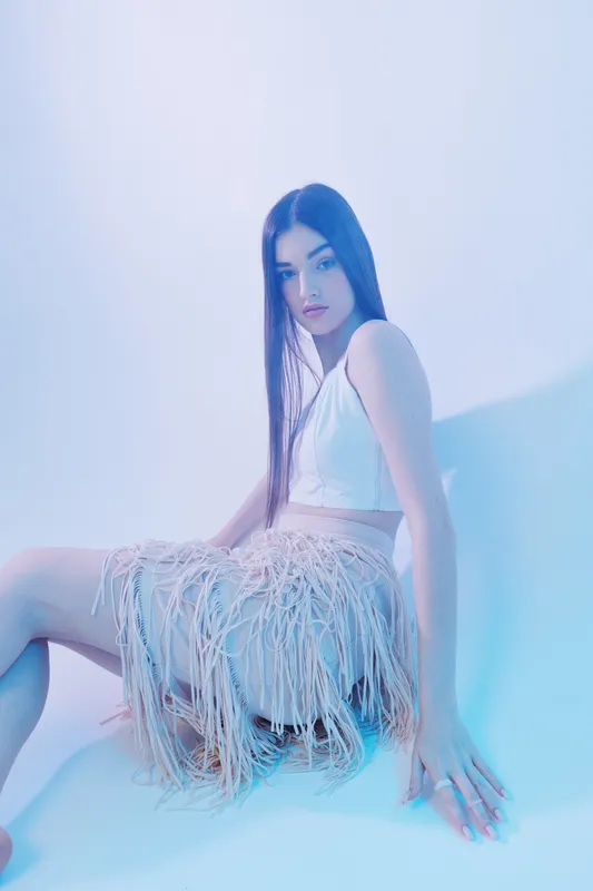 Young woman beauty fashion model poses sitting in a white top and fringe skirt against a soft blue background. Concept of style, elegance, and modern trends.