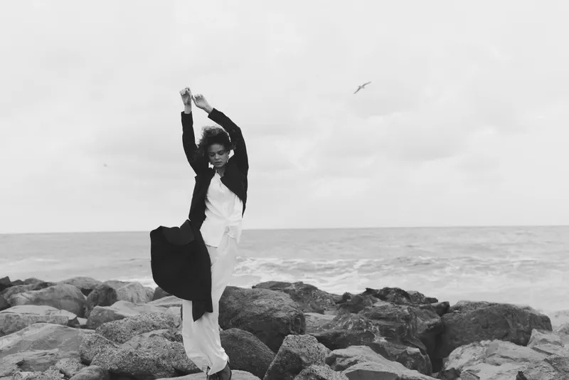Dancer on rugged rocks by the sea lifts arms in a dynamic pose, windswept silhouette against a gray sky, capturing movement, balance and grace in a black and white coastal scene