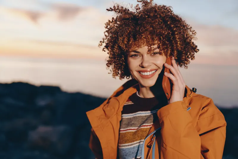 Woman lifestyle outdoors, smiling, curly hair in sunset glow, orange jacket, holding phone to ear, mountain landscape behind, warm natural light, relaxed cheerful mood