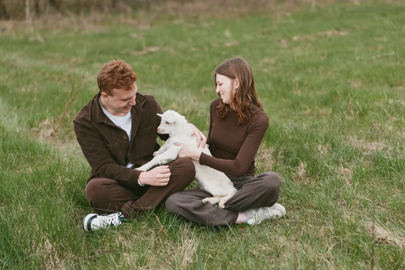 Cheerful couple outdoors in a grassy field with a playful goat kid, warm sunlight, candid moment, relaxed posture, natural colors, companionship and joy in a tranquil rural setting