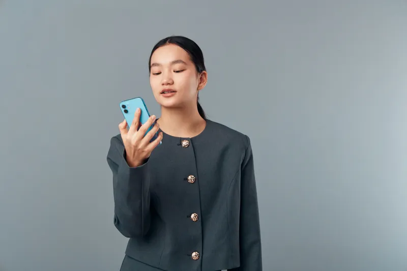 Young woman with closed eyes holding smartphone in hand for communication and social media. Female portrait in formal jacket with modern device on gray background for technology use.