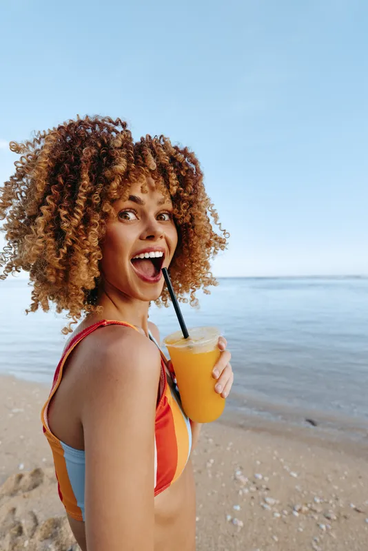 Woman curly hair beach cocktail straw summer happiness vacation drinking smiling portrait by ocean sunlight she holds an orange cocktail with straw, laughing and relaxing on sandy shore