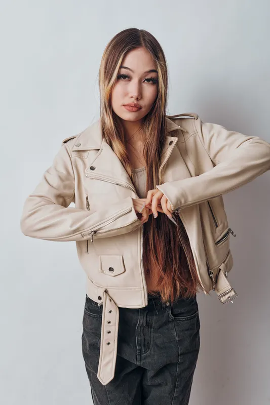 Young woman standing in light beige leather jacket with long straight hair unzipping sleeve zipper against plain white background in casual fashion pose.