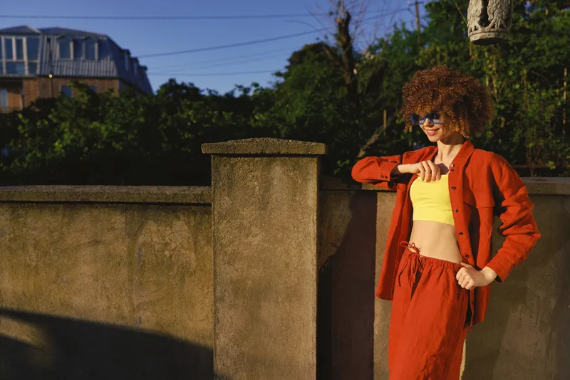 A stylish woman outdoors in a bright orange casual outfit checks a smartwatch against a stone barrier, radiating confidence. A lifestyle moment in sunny fashion