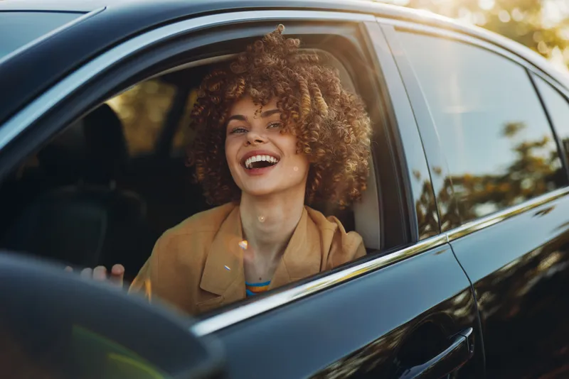Curly haired woman smiling inside a car, sunlight streaming through the window, capturing a candid lifestyle moment that conveys joy, warmth and confidence