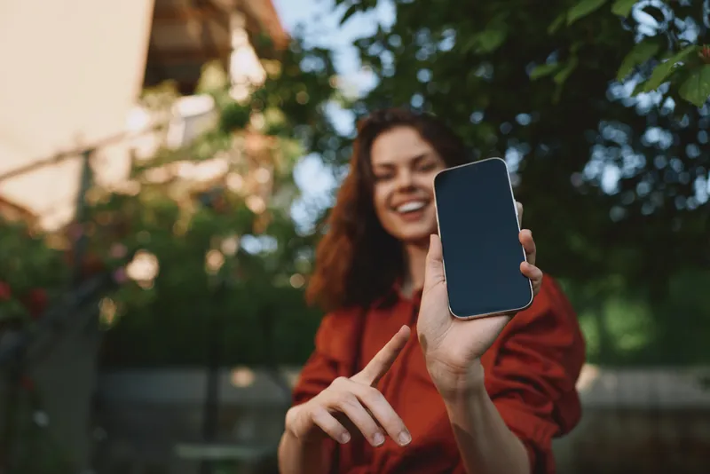 Young woman takes a carefree outdoor selfie with a smartphone, smiling at the camera as lush greenery, warm sunlight, and a blurred background create a relaxed, candid lifestyle moment