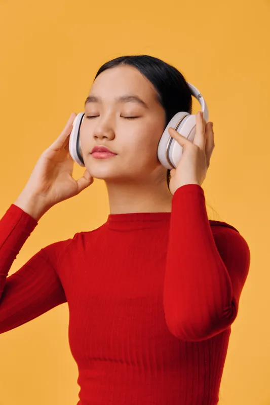 Young woman in red sweater with closed eyes enjoying music wearing white wireless headphones on orange background for relaxation and technology concept.