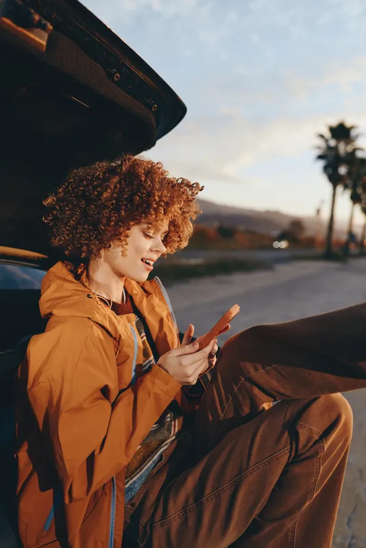 Woman lifestyle roadtrip, she sits near car trunk with open suitcase, wearing orange jacket, making peace sign, smiling outdoors, curly hair, warm sunlight, autumn