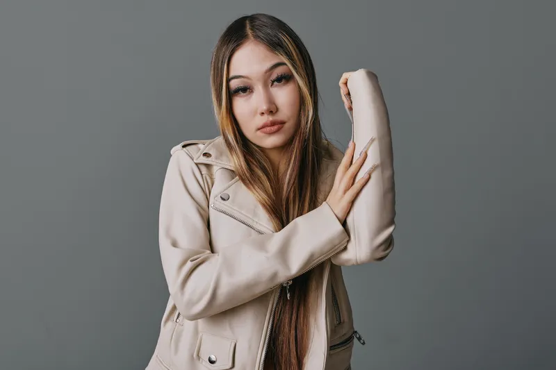 Young woman with long straight hair wearing beige jacket posing with raised arm against gray background, looking at camera with calm and confident expression in studio portrait.