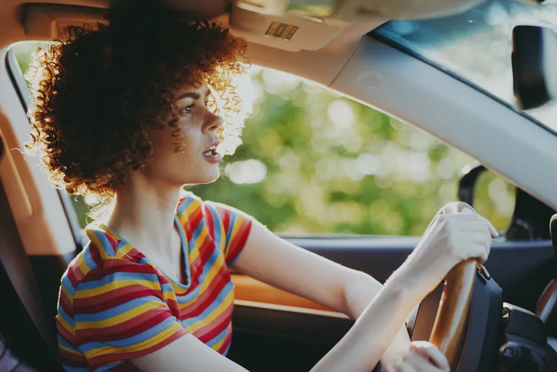 Curly-haired woman driving a car wearing a striped shirt, hands on the wheel. Bright sunlight through the window creates a warm retro mood. Natural light highlights textures, candid moment,