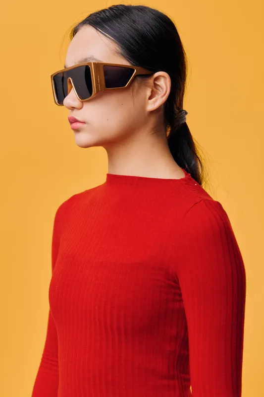 Young woman in red sweater wearing large sunglasses with black lenses and brown frame standing against yellow background with serious expression in profile view.