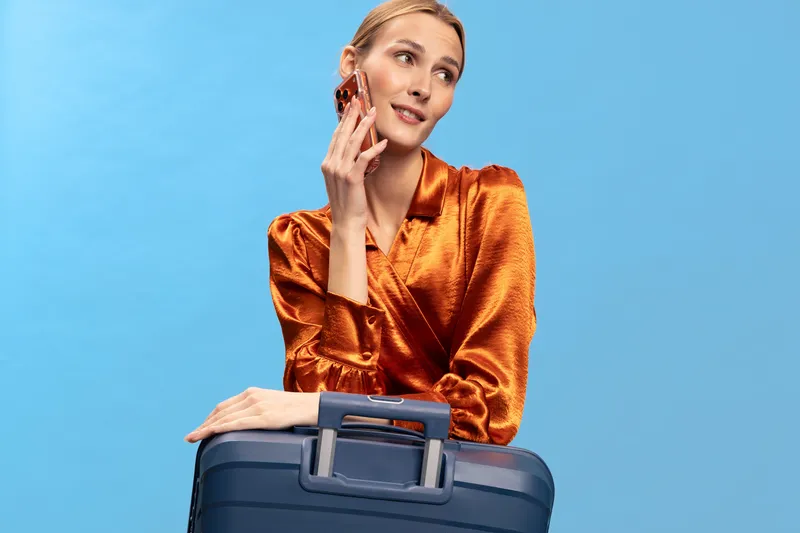 Young woman with blonde hair in shiny orange blouse holding blue suitcase and smiling while looking away on blue background. Concept of travel, vacation, and lifestyle with positive mood.