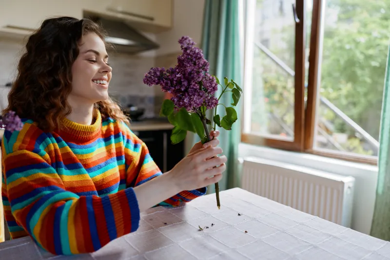 A cheerful woman sits at a dining table in a bright home, admiring a bouquet of lilac flowers, wearing a multicolored striped sweater, creating a cozy lifestyle moment