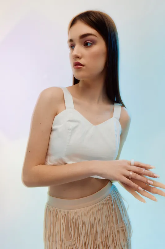 Young woman with long brown hair wearing a white top and beige skirt with fringes looks to the side. Studio shot with soft lighting. Concept of woman beauty fashion and style.