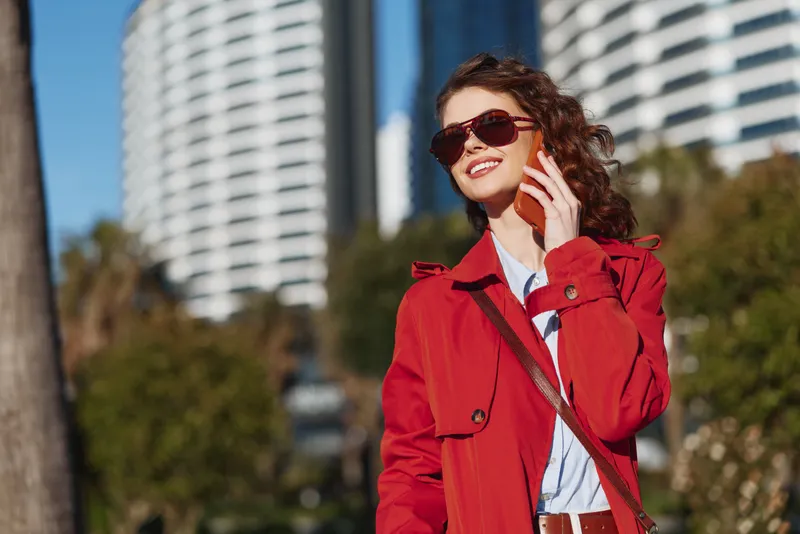 Stylish woman outdoors wearing a red coat and sunglasses, talking on a phone in a bright urban setting with modern buildings, capturing a confident candid moment