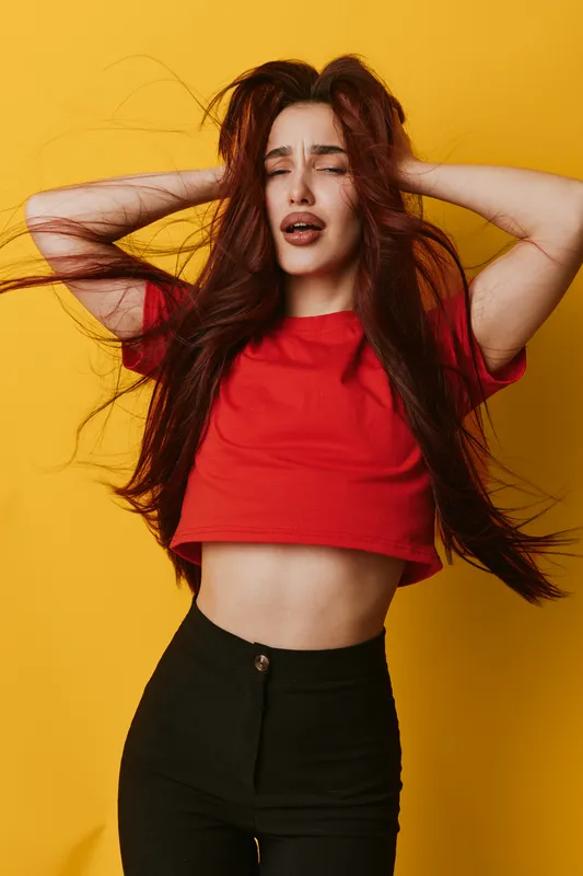 Woman with long hair wearing red crop top and black pants posing with eyes closed hands on head against yellow background. Fashion, beauty, casual style and emotion in studio portrait.