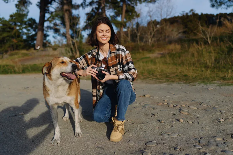Dog and woman posing outdoors on a sandy beach, smiling with a camera in hand, casual plaid shirt and boots, natural scenery and relaxed lifestyle
