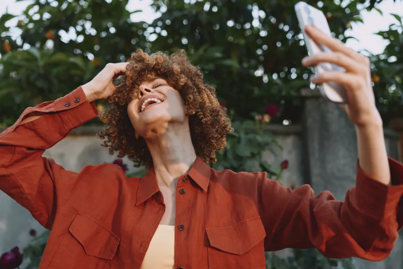 Smiling woman with curly hair taking a selfie outdoors, wearing a red shirt. Casual lifestyle portrait with natural light and warm mood, celebrating confidence, youth, and self expression