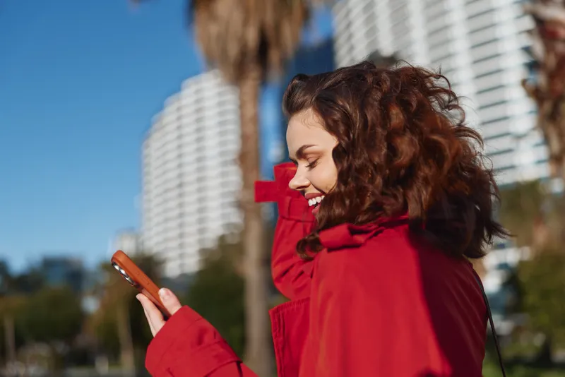 Bright portrait of a smiling woman in a red coat checking her smartphone outdoors, with urban buildings and palm trees in the background under sunny weather.