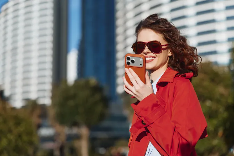 Smiling woman in a red coat takes a selfie with a smartphone in an urban outdoor setting, tall buildings in the background, bright mood with clear sun.