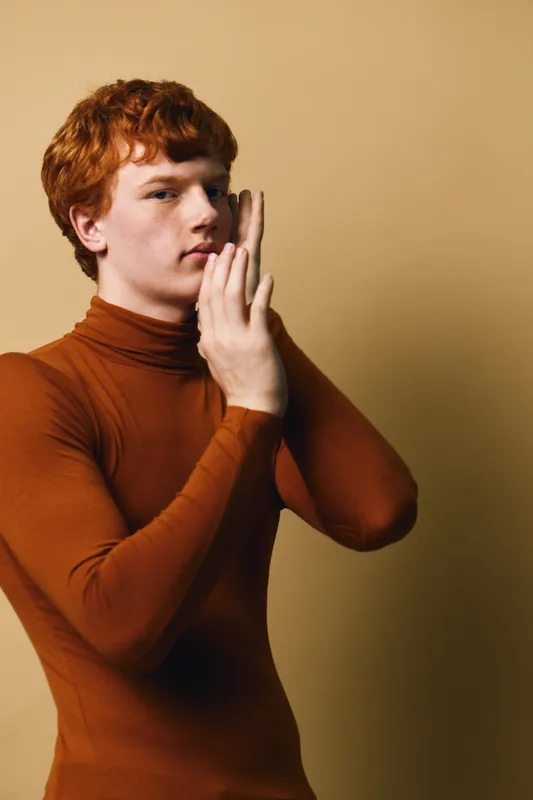 Young man with red hair in brown turtleneck posing with hands near face against beige background, studio portrait, fashion and minimalism concept, thoughtful expression