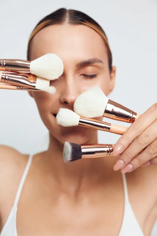 Young woman holding multiple makeup brushes near her face with closed eyes, demonstrating beauty tools on a white background. Concept of cosmetics, skincare, and makeup routine.