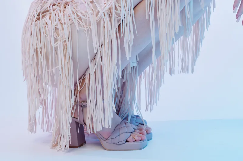 Close-up of a woman in fashionable fringed clothing wearing stylish high-heeled sandals. The image highlights beauty, fashion, and elegance in a minimalist studio setting.