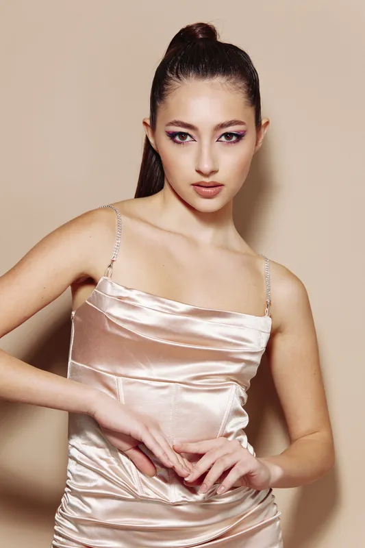 Young woman with glossy hair in sleek light dress poses confidently in studio. Bold purple eye makeup adds artistic flair for beauty, fashion, and creative concept.