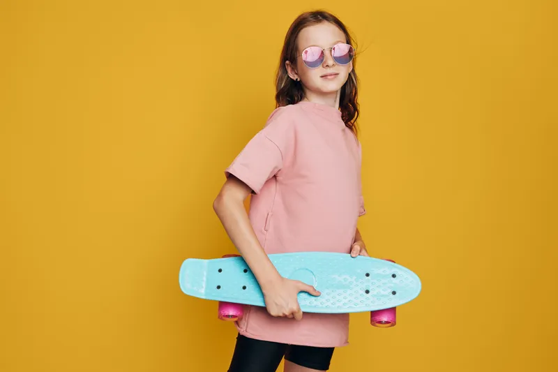 Young girl in pink oversized t-shirt holding blue skateboard against yellow background. She wears reflective sunglasses and stands confidently. Concept of youth, leisure, and urban lifestyle.