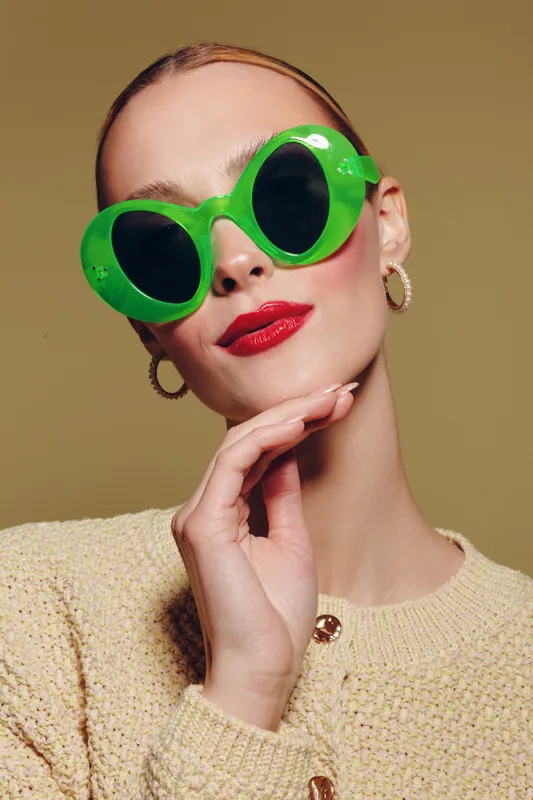 Stylish woman wearing large green sunglasses and red lipstick poses with hand near chin against beige background. Fashion portrait with trendy accessories and confident expression.