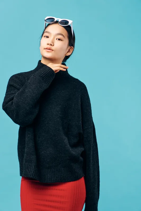 Young woman posing in black sweater and red skirt with sunglasses on head against blue background. Model with calm expression standing in casual stylish outfit and long sleeves.
