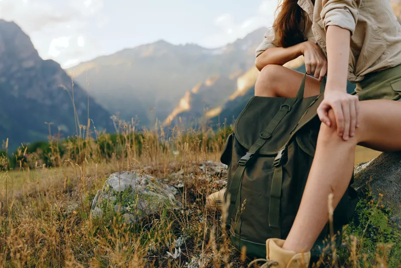 Young woman traveler with backpack resting on rock in mountain field holding hand of companion during outdoor adventure in sunny nature landscape.