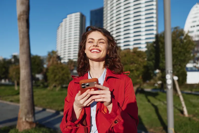 Young woman in red coat smiles holding smartphone outdoors with modern buildings and trees in background on sunny day in city park.