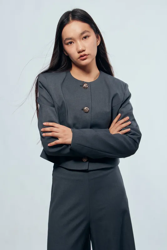 Young woman with long dark hair wearing grey business suit standing with crossed arms against light background. Serious facial expression for professional portrait or fashion.