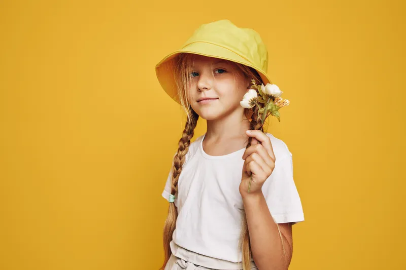 Young girl with braided hair wearing a yellow hat and white shirt holds small wildflowers on a yellow background. Studio portrait with natural expression and soft lighting.