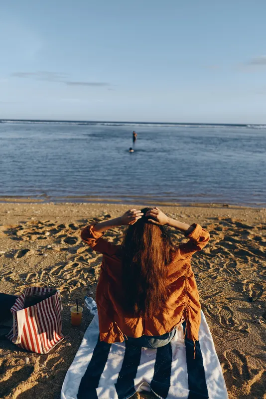 A person sits on a striped towel on a sandy beach, facing calm water with hands on head, conveying stress, contemplation, and emotional fatigue by the sea.