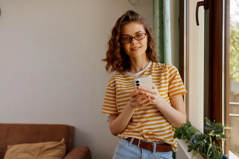Casual woman with curly hair standing by a window using a smartphone in a bright home setting, striped shirt, relaxed lifestyle, everyday tech use, warm natural light.