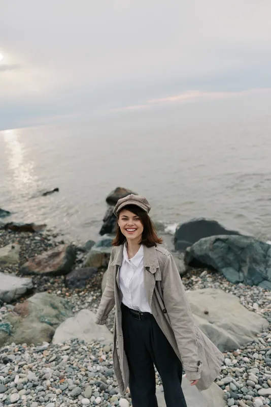 Happy young woman in casual outfit enjoys a pebble beach near calm sea at cloudy weather. She smiles and wears a hat with a long coat, casual style outdoors with natural background.