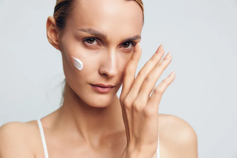 Young woman applying face cream near cheek, gentle beauty routine, skincare cosmetics care on clean background isolated, hand gesture showing moisturize texture, hygiene spa wellness