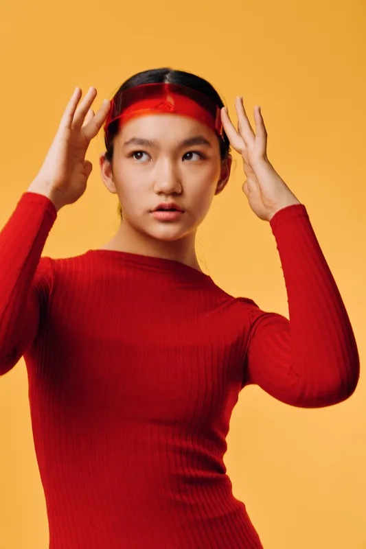 Young woman in red sweater adjusting red transparent visor on head looking sideways on orange background. Female fashion model with sleek hair wearing casual modern outfit in studio light.