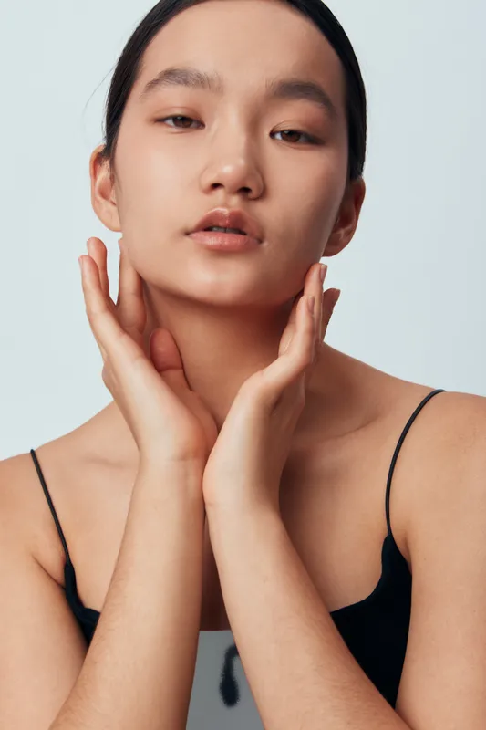 Young woman with clear skin and black hair poses with hands near face wearing black strap top studio portrait. Beauty, skincare, elegance, confident model with natural makeup and minimalistic style.